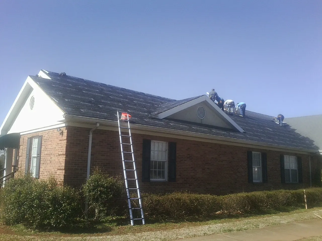 Crew tearing shingles off a brick ranch roof with a ladder set — Emergency Roof Repair work by our crew in De Ruyter