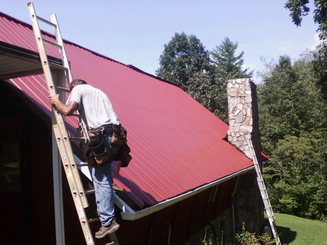 Roofer on a ladder beside a red metal roof and stone chimney — Soffit & Fascia Repair by Roofing Brothers in De Ruyter